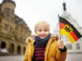Uśmiechnięty chłopiec z blond włosami trzyma flagę Niemiec. W tle widać zabytkowe budynki, przypominające te, które można spotkać w niemieckich miasteczkach, gdzie znajdują się szkoły.