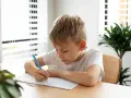 Chłopiec pisze w zeszycie niebieskim długopisem przy biurku.