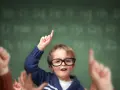 Chłopiec w okularach, jak przewodniczący klasy, podnosi rękę z palcem do góry, gotowy do odpowiedzi. W tle widać litery alfabetu na tablicy.