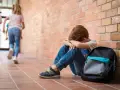 Chłopiec z fobią społeczną u dzieci siedzi przy ceglanej ścianie, podczas gdy inni biegają obok.