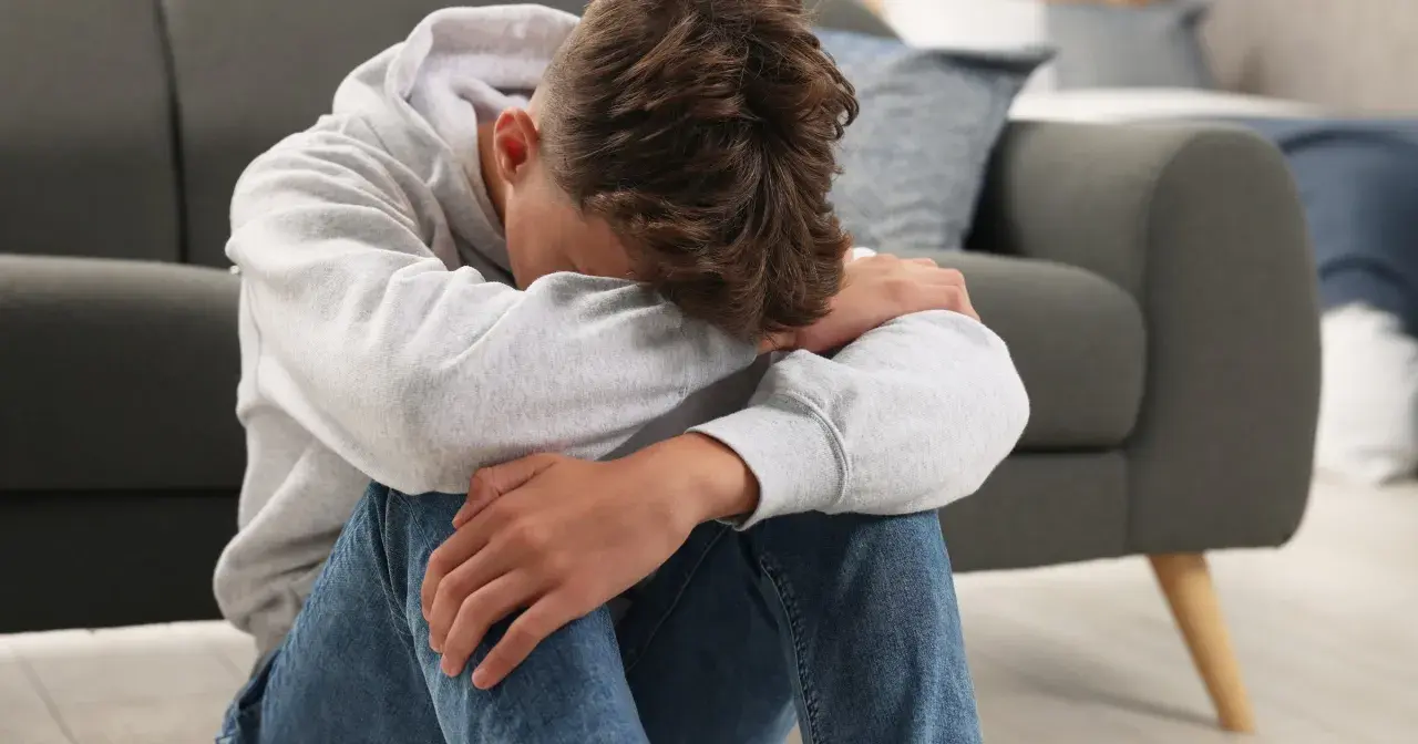 Chłopiec w szarej bluzie z kapturem siedzi na podłodze, przytulając kolana. Wygląda na smutnego, może potrzebuje rozmowy z psychologiem bez zgody rodzica.