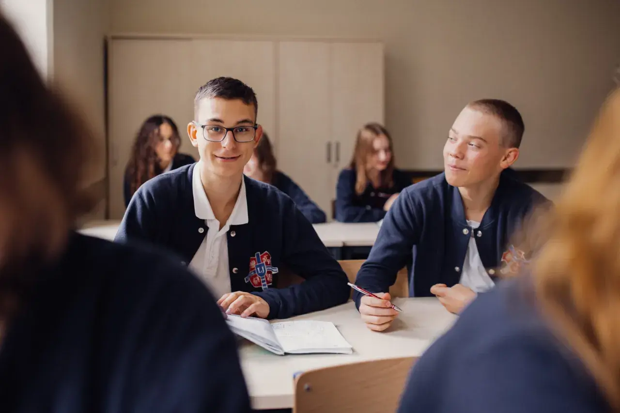 Uczniowie liceum podczas lekcji. Chłopiec w okularach uśmiecha się, a jego kolega pisze w zeszycie. To obraz nauczania domowego liceum.