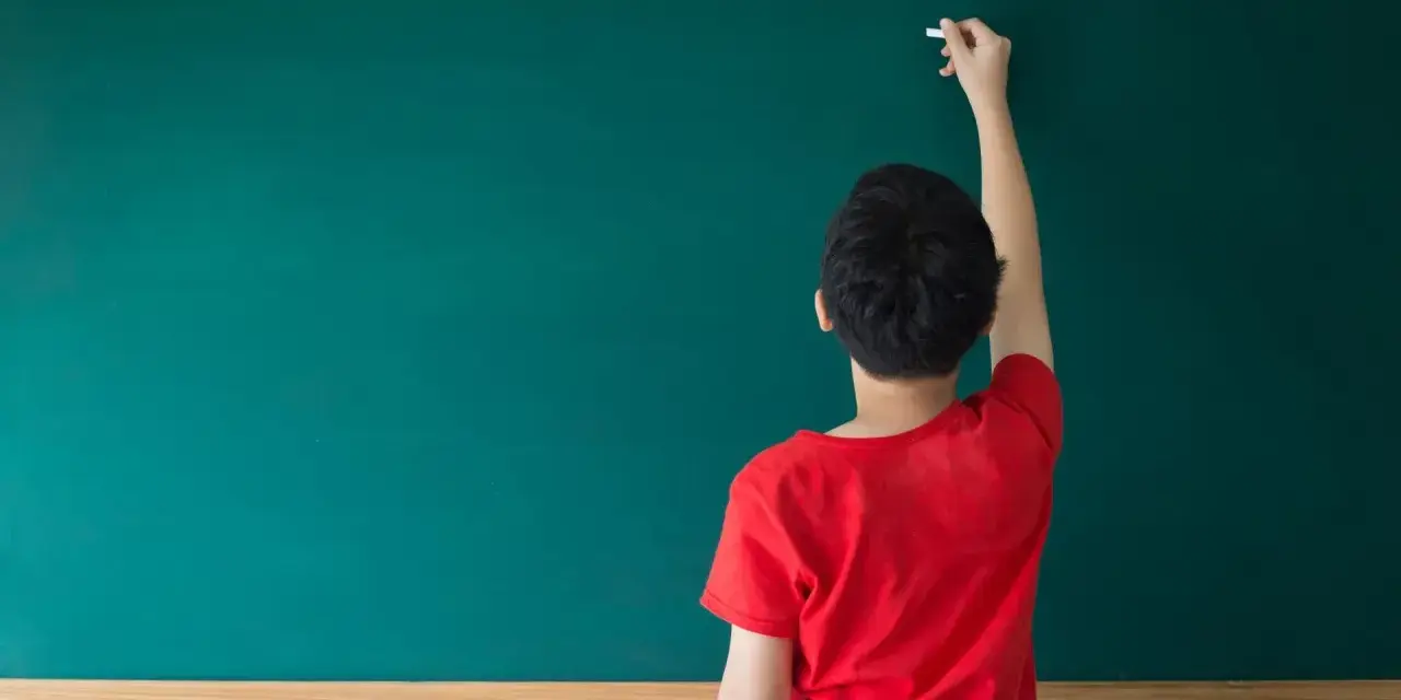Chłopiec w czerwonej koszulce pisze kredą na zielonej tablicy. Zastanawia się, czy przed "może" stawiamy przecinek.