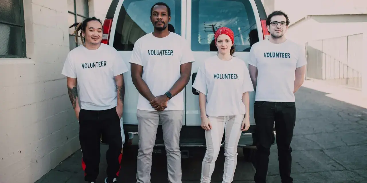 Czterech wolontariuszy w białych koszulkach z napisem "VOLUNTEER" stoi przed vanem. To może być wzór zgody rodzica na wolontariat.