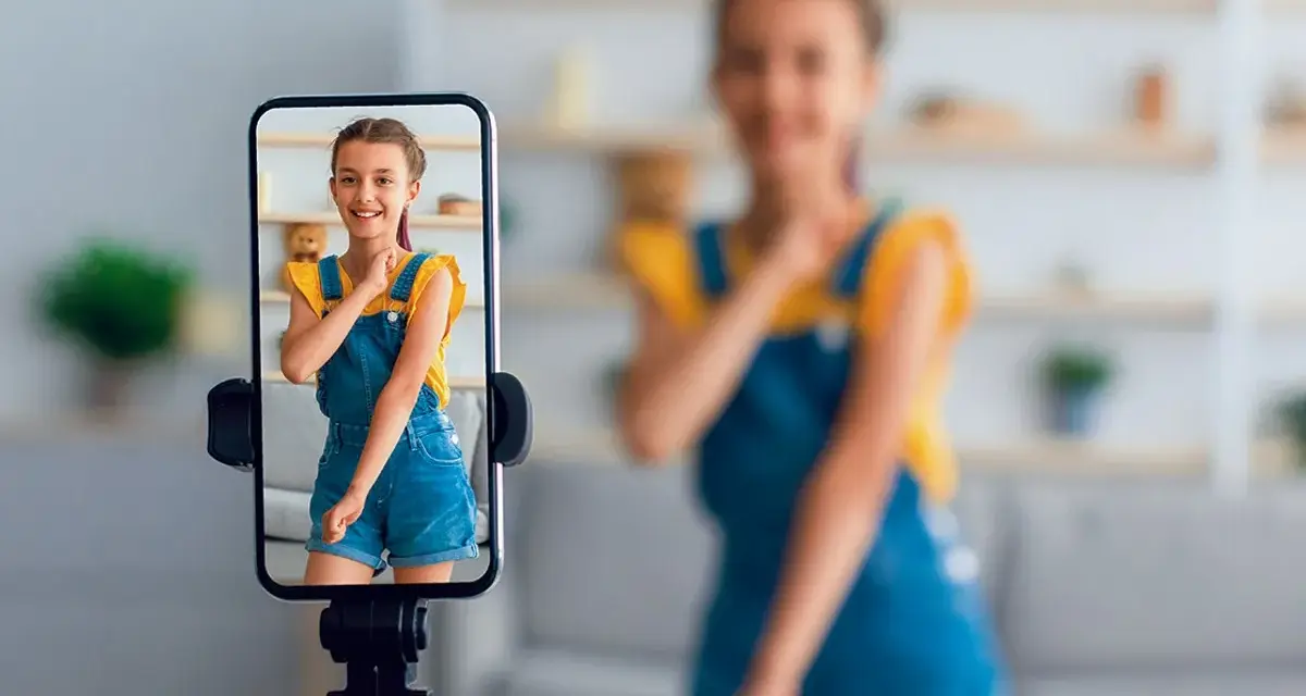 Dziewczynka w ogrodniczkach na ekranie telefonu, nagrywana przez mamę. To przykład zjawiska sharenting co to znaczy dzielenie się życiem dziecka w sieci.