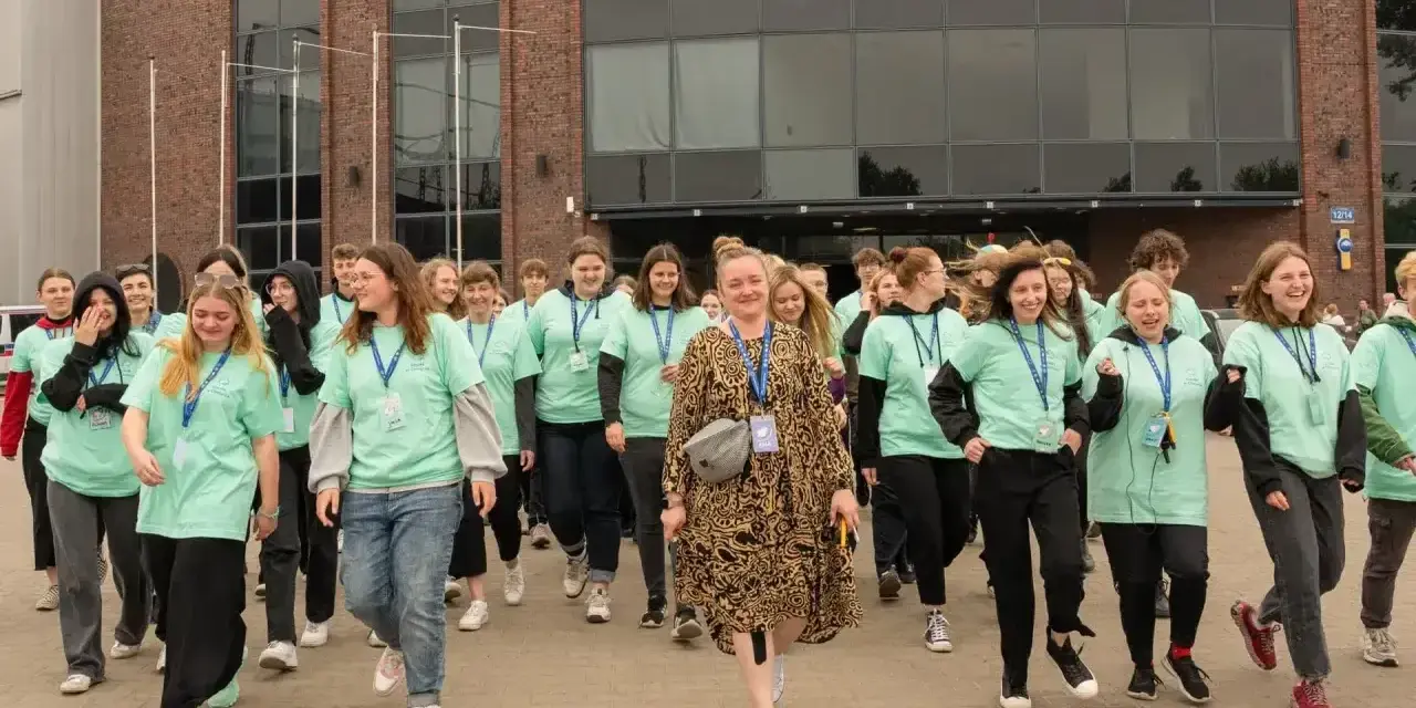Grupa młodych ludzi w zielonych koszulkach idzie przed budynkiem. Jak się dostać do szkoły w chmurze? Zapytaj ich!