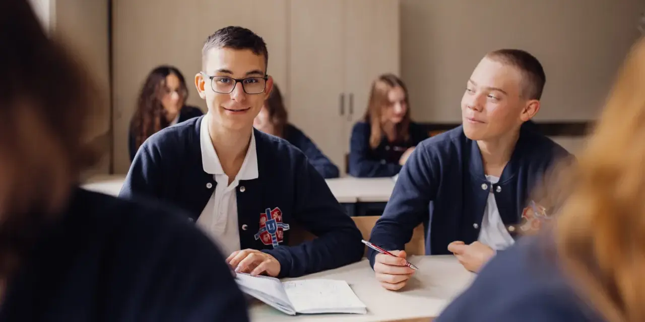 Uczniowie liceum podczas lekcji. Chłopiec w okularach uśmiecha się, a jego kolega pisze w zeszycie. To obraz nauczania domowego liceum.