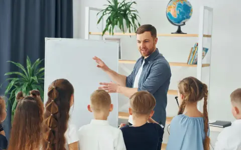 Nauczyciel tłumaczy coś dzieciom przy tablicy. To mogą być dni dyrektorskie, podczas których uczniowie poznają szkołę.