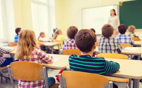 Dzieci siedzą w ławkach podczas lekcji religii w szkole, słuchając nauczycielki stojącej przy tablicy.