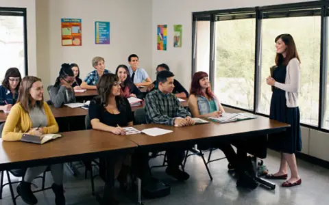 Uczniowie słuchają nauczycielki podczas lekcji. W klasie panuje atmosfera skupienia, jak podczas ważnej rady pedagogicznej.
