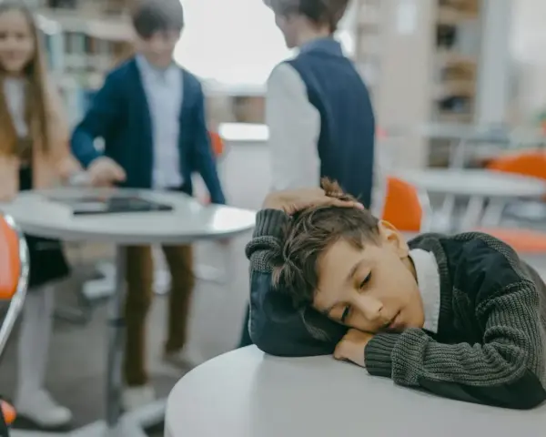 Samotne dziecko w klasie, zmęczone lekcją, opiera głowę na stole. W tle inni uczniowie.