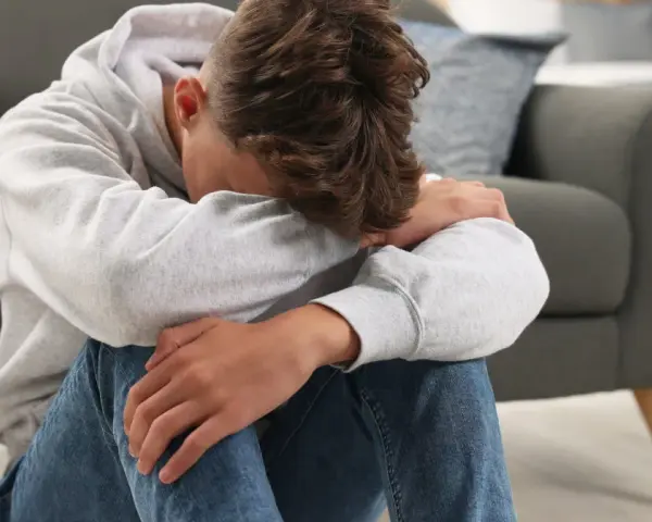 Chłopiec w szarej bluzie z kapturem siedzi na podłodze, przytulając kolana. Wygląda na smutnego, może potrzebuje rozmowy z psychologiem bez zgody rodzica.