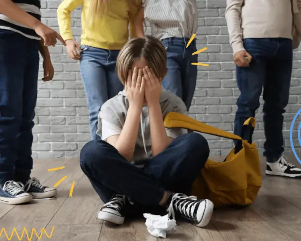 Dziecko zakrywa twarz, otoczone przez inne dzieci, które wskazują palcami. To obraz tego, czym jest bullying.