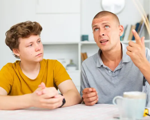 Ojciec tłumaczy synowi, jak rozmawiać z nastolatkiem. Chłopak słucha uważnie, a mężczyzna gestykuluje, podkreślając swoje słowa.