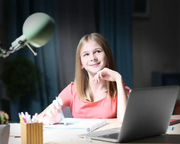 Uśmiechnięta dziewczynka z długopisem w dłoni, zastanawia się nad zadaniem. Obok laptop, dlatego jaka to część mowy?