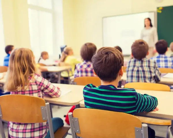 Dzieci siedzą w ławkach podczas lekcji religii w szkole, słuchając nauczycielki stojącej przy tablicy.