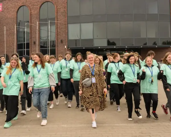 Grupa młodych ludzi w zielonych koszulkach idzie przed budynkiem. Jak się dostać do szkoły w chmurze? Zapytaj ich!