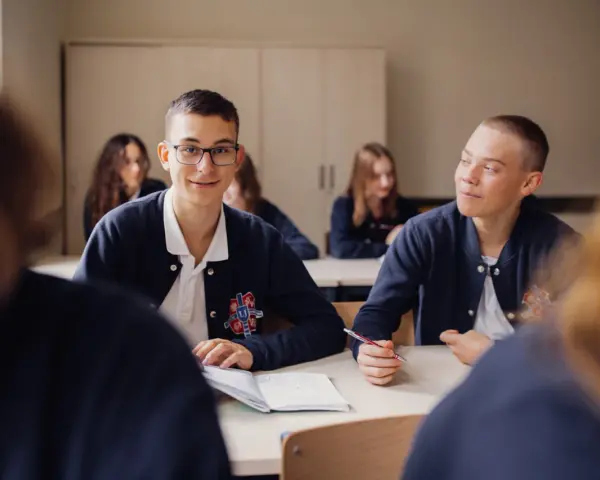 Uczniowie liceum podczas lekcji. Chłopiec w okularach uśmiecha się, a jego kolega pisze w zeszycie. To obraz nauczania domowego liceum.