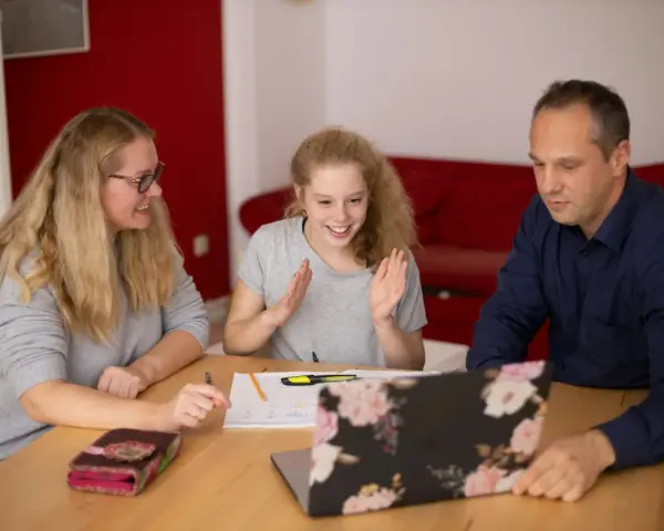 Rodzina przy stole uczy się zdalnie. Córka z entuzjazmem tłumaczy coś rodzicom, szukając sposobu, jak przekonać ich do swoich racji.