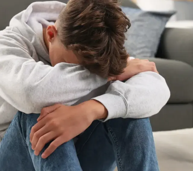 Chłopiec w szarej bluzie z kapturem siedzi na podłodze, przytulając kolana. Wygląda na smutnego, może potrzebuje rozmowy z psychologiem bez zgody rodzica.