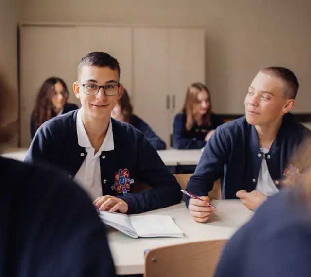 Uczniowie liceum podczas lekcji. Chłopiec w okularach uśmiecha się, a jego kolega pisze w zeszycie. To obraz nauczania domowego liceum.