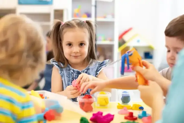 Uśmiechnięta dziewczynka z kitkami bawi się plasteliną, zastanawiając się, w jakim wieku do szkoły pójdzie. Dzieci tworzą kolorowe figurki.