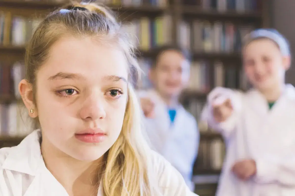 Smutna dziewczynka z łzami w oczach, podczas gdy dwie inne postacie w białych fartuchach wskazują na nią palcami. To obraz tego, czym jest bullying.