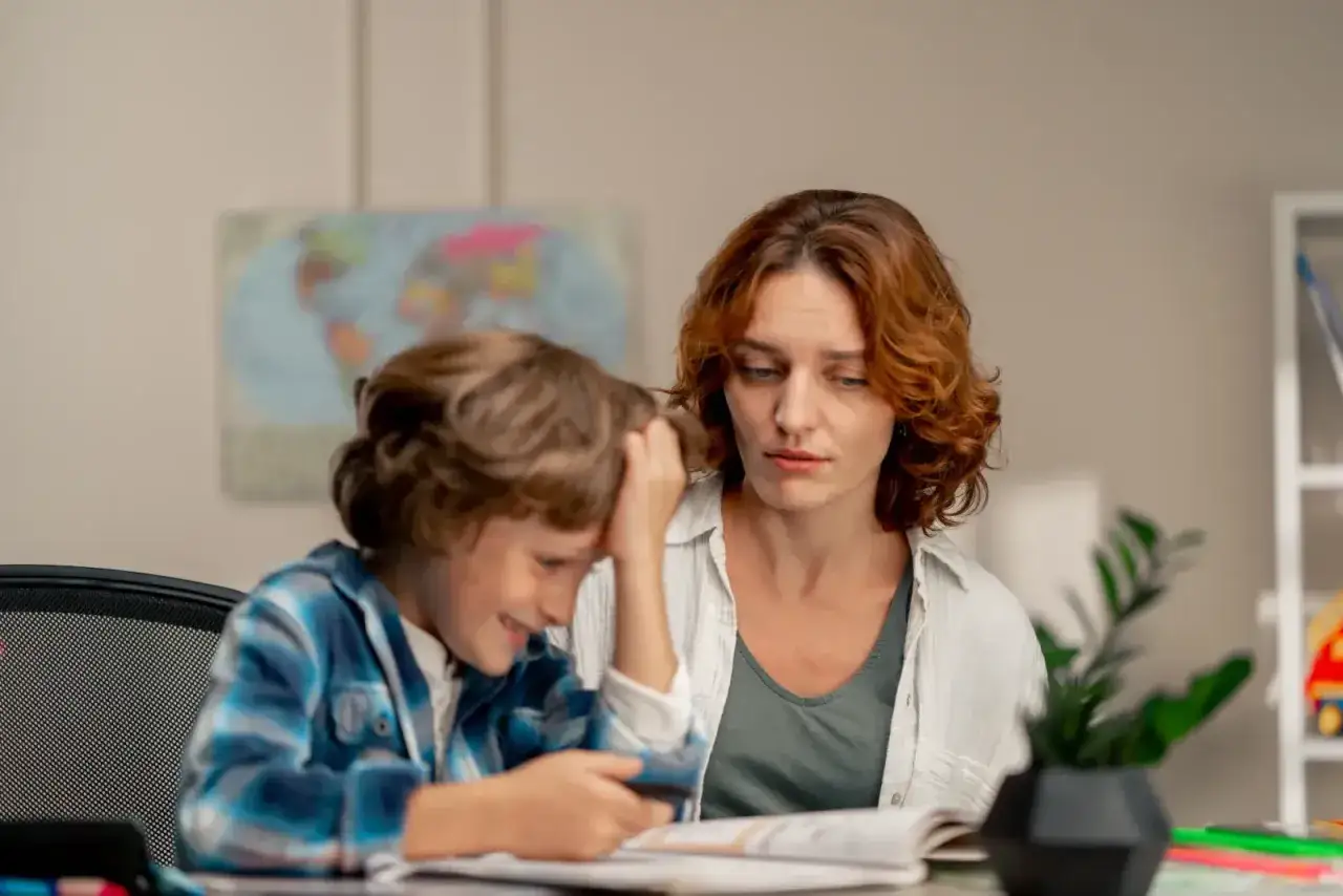 Mama pomaga synkowi w odrabianiu prac domowych. Chłopiec z frustracją drapie się po głowie, podczas gdy mama patrzy na niego z troską.