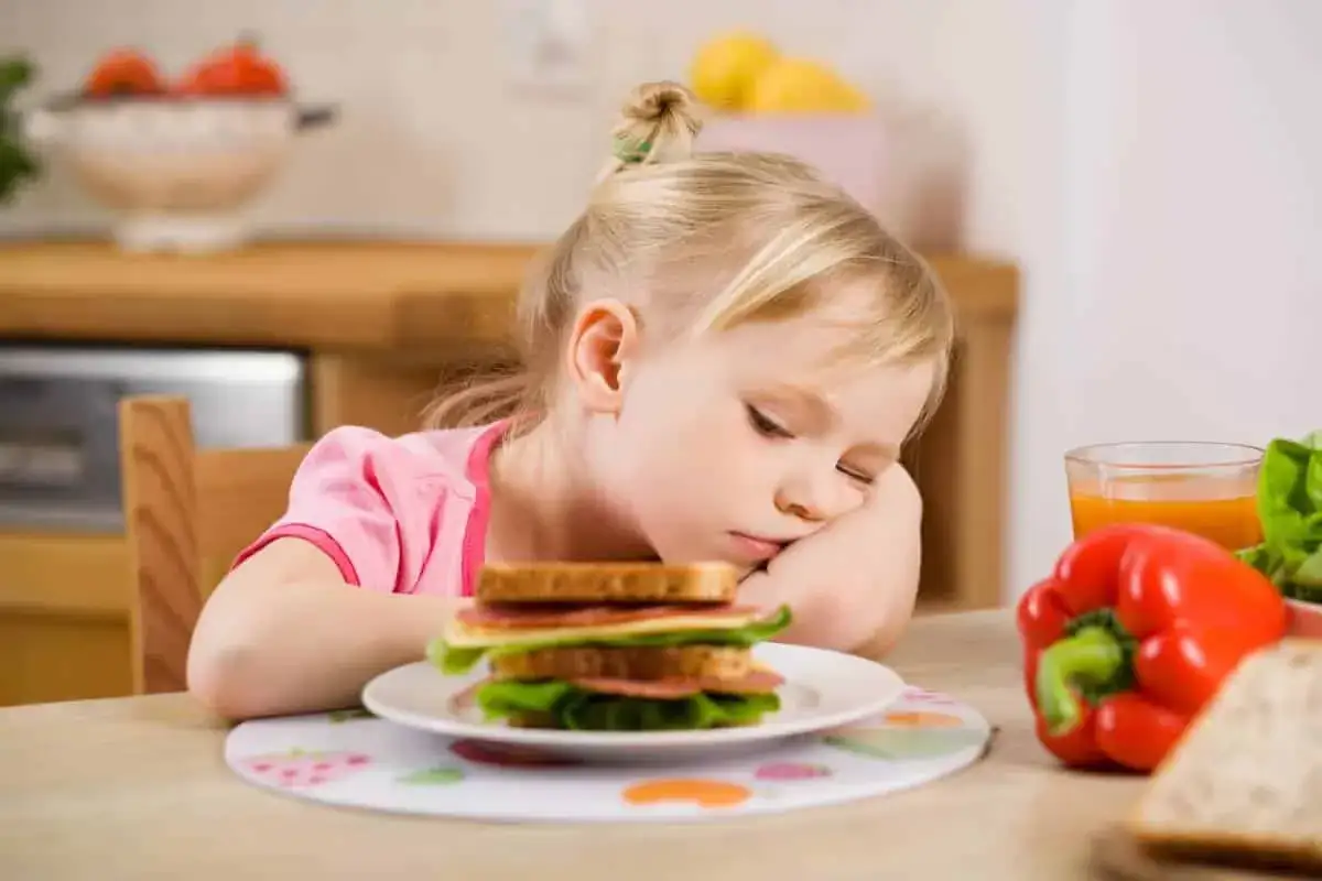 Smutna dziewczynka z zaburzeniami odżywiania u dzieci patrzy na kanapkę. Obok sok i warzywa.